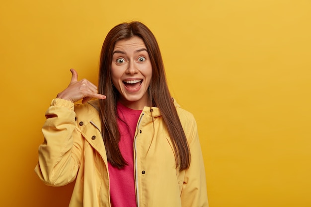 Body language concept. Positive brunette woman makes call gesture, says call me back, wears yellow anorak, asks for number, looks gladfully at camera