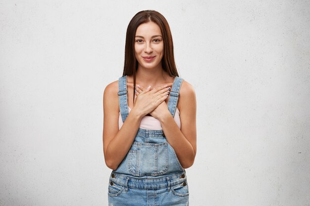 Body language. Gorgeous friendly looking young female in denim jumpsuit with kind big heart and beaming smile, holding hands crossed on her chest, expressing gratefulness and gratitude, saying Thanks