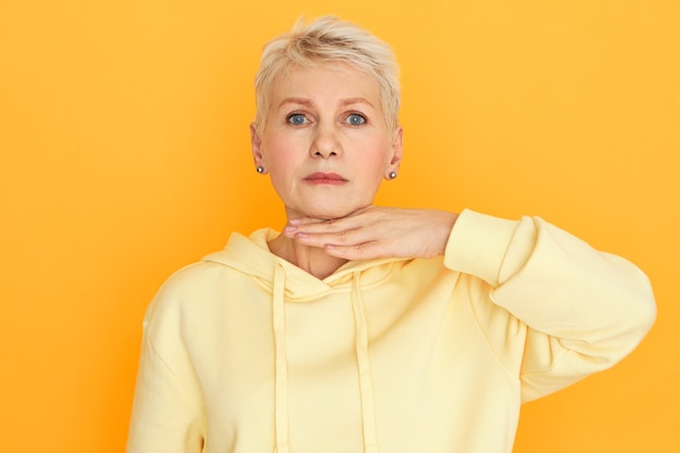 Body language. Horizontal studio image of frustrated angry woman pensioner in stylish hoodie holding palm at her neck, making threatening gesture, menacing you, having displeased tired look