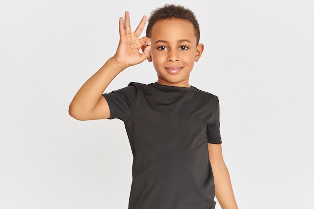 Free photo body language. portrait of friendly looking positive dark skinned little boy in t-shirt connecting fore finger and thumb making approval gesture, showing okay sign, saying everything is fine