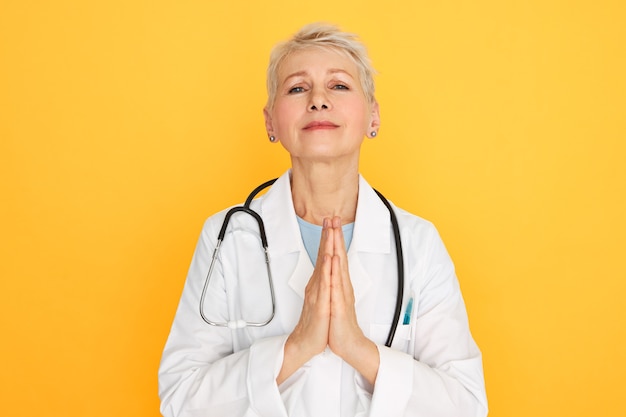 Free photo body language. portrait of sad unhappy middle aged female surgeon in medical unifrom having mournful look, pressing hands together, praying, hoping that terminally ill patient will recover