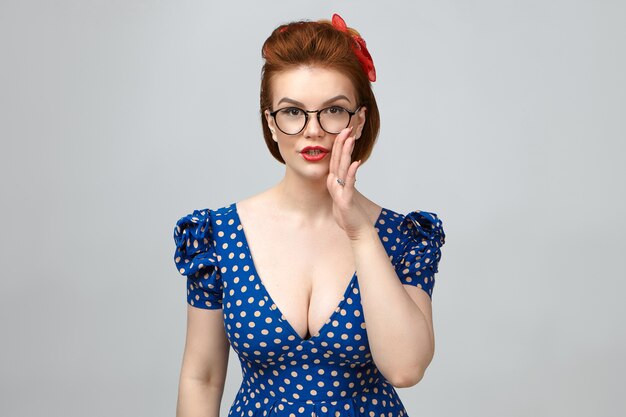 Body language. Pretty young European woman wearing glasses, elegant dress and bright make up holding hand at her mouth, sharing top secret or confidential information with you, having mysterious look