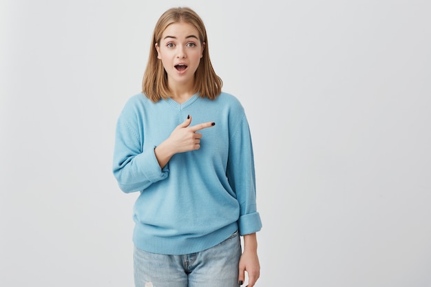 Free photo body language. shocked, excited, surprised girl with straight fair hair and opened mouth, bugged eyes, pointing with index finger at copy space for your promotional text or advertisment