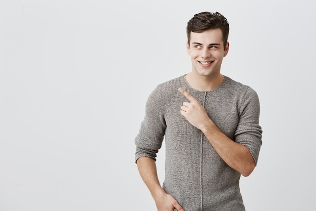 Body language. Shocked, excited, surprised guy with dark hair, broadly smiling with teeth, dressed casually, pointing with index finger at copy space for your promotional text or advertisment