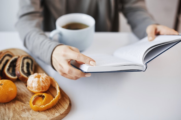 Book is like chamber of knowledge. Smart modern female reading favorite novel during breakfast, enjoying drinking hot tea in calm and cozy atmosphere, peeling tangerine and eating