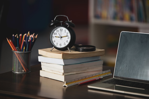 Book, laptop, pencil, clock on wooden table in library, education learning concept