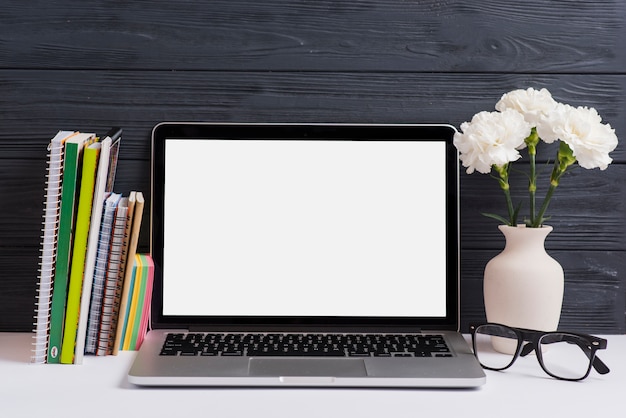 Books; adhesive notes; laptop; eyeglasses and vase on white desk against wooden wall