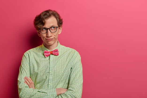 Free photo bored adult man geek sighs and looks thoughfully aside, keeps arms folded, wears spectacles and elegant clothes, thinks over future plans, stands against rosy wall, blank space on right side.