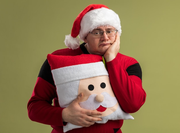 Bored adult man wearing glasses and santa hat holding santa claus pillow keeping hand on face looking at side isolated on olive green wall