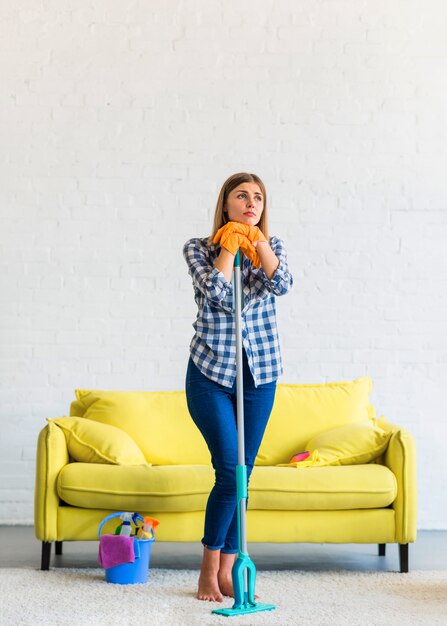 Bored contemplated young woman holding mop standing in the living room