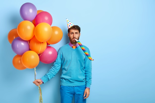 Free photo bored guy with birthday hat and balloons posing in blue sweater