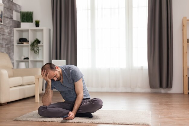 Bored man sitting on carpet in living room browsing on smartphone during quarantine.