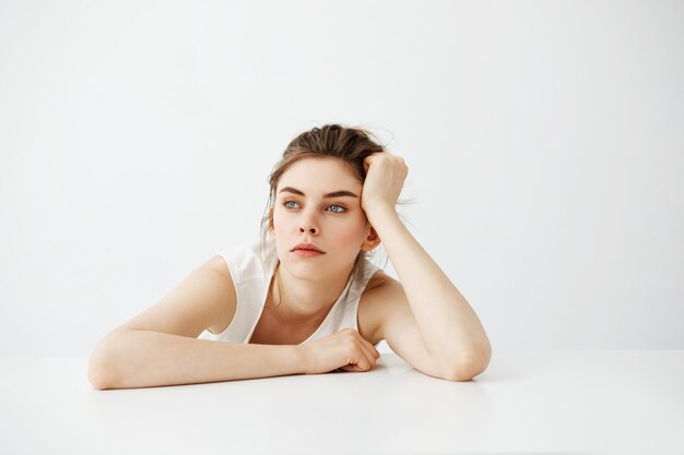 Bored tired young pretty woman with bun thinking dreaming sitting at table over white background.