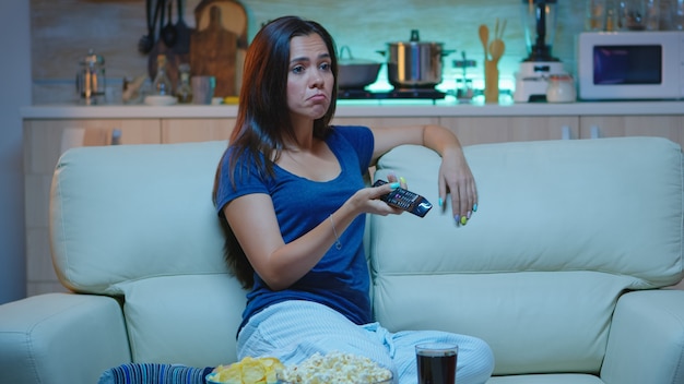 Free photo bored woman resting watching tv in living room sitting on couch. tired from work, home alone late at night lady relaxing on sofa in front television holding remote control choosing channel with movie