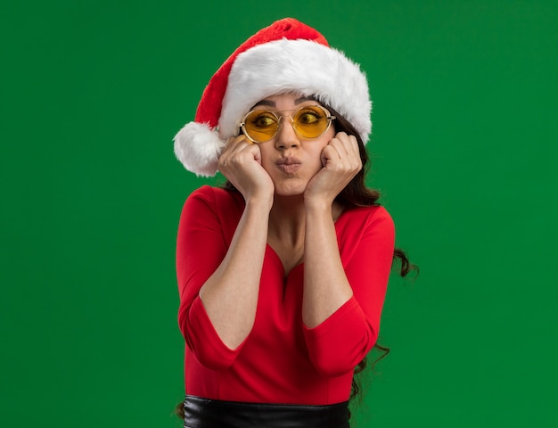 Free Photo bored young pretty girl wearing santa hat and glasses keeping hands on chin puffing cheeks looking at side isolated on green background with copy space