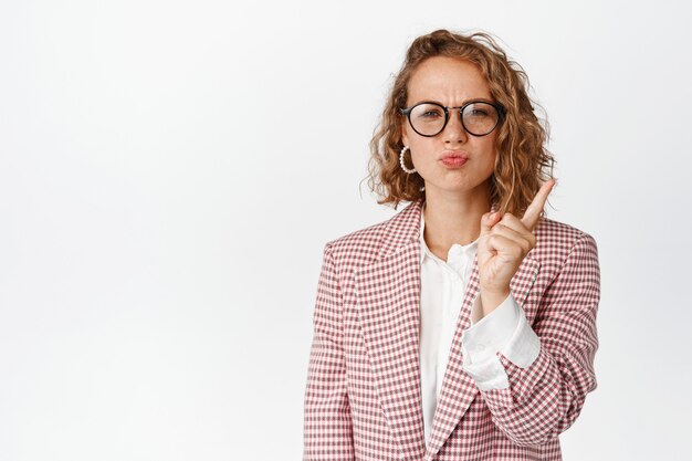 Bossy business woman in glasses scolding someone, shaking finger in disapproval, boss around, standing on white