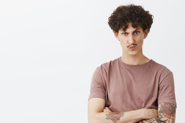 Bossy displeased and suspicious good-looking stylish jewish guy with curly hair and moustache in brown t-shirt holding hands crossed on chest smirking and frowning doubtful over gray wall