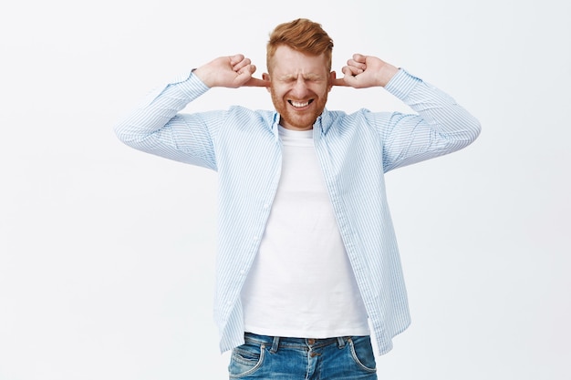 Free photo bothered and annoyed redhead caucasian man feeling discomfort hearing loud noise or serene, covering ears with index fingers like earplugs, squinting and clenching teeth from dislike