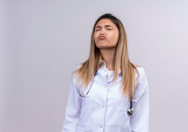 Bothered young beautiful woman doctor wearing white coat with stethoscope blowing cheeks