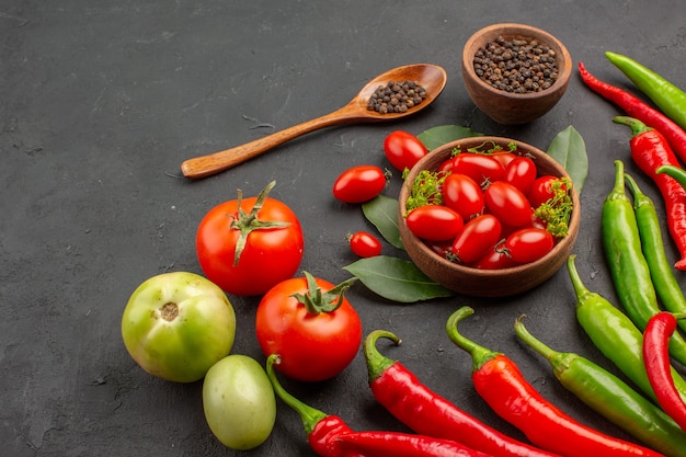 Bottom close view a bowl of cherry tomatoes hot red and green peppers and tomatoes black pepper in a wooden spoon a bowl of black pepper on black background