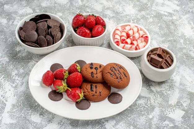 Bottom close view cookies strawberries and round chocolates on the white oval plate surrounded bowls with candies strawberries and chocolates on the ground with copy space