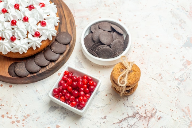 Bottom half view cake with white cream on cutting board bowls with berries and chocolate cookies tied with rope on light grey table