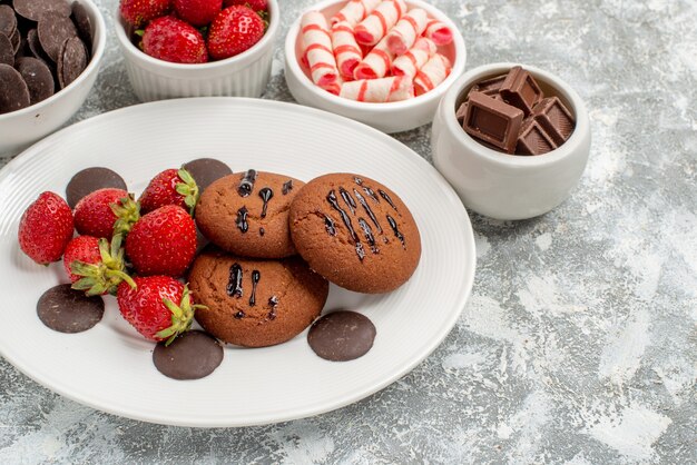 Bottom half view cookies strawberries and round chocolates on the white oval plate surrounded bowls with candies strawberries and chocolates on the grey-white table