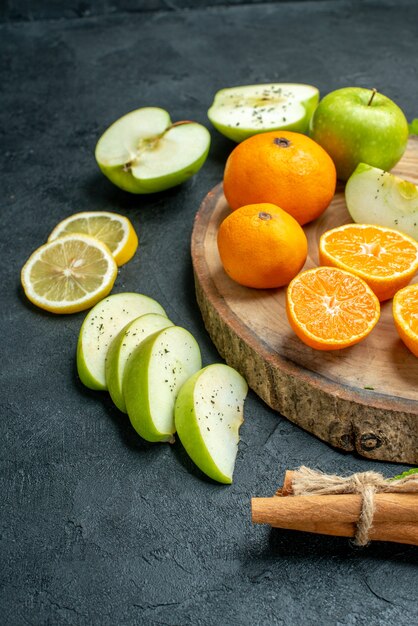 Bottom half view cut apples and mandarines on round wood board cinnamons tied with rope lemon slices on dark table