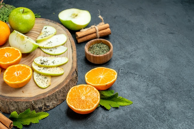 Bottom half view fresh apple and orange slices on wood board dried mint powder in bowl cinnamon sticks pine tree branches on black table free space