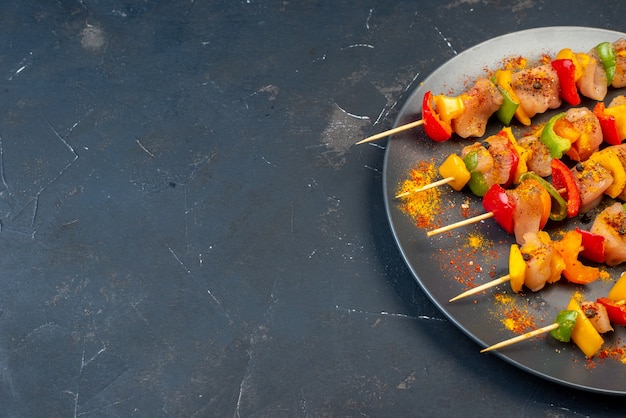 Bottom half view raw chicken skewers with spices on wooden board on dark table with copy space