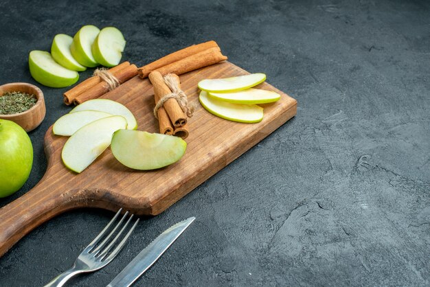 Bottom view apple slices and cinnamon sticks on chopping board knife and fork dried mint powder in small bowl on dark table with free space