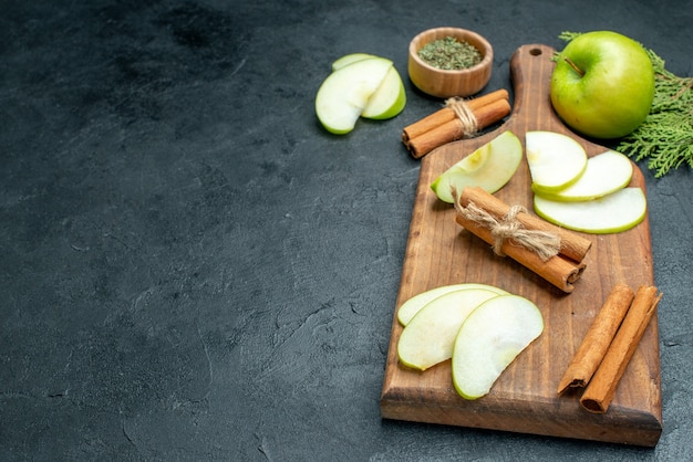 Bottom view apple slices and cinnamon sticks on wood serving board dried mint powder in small bowl on dark table with copy place