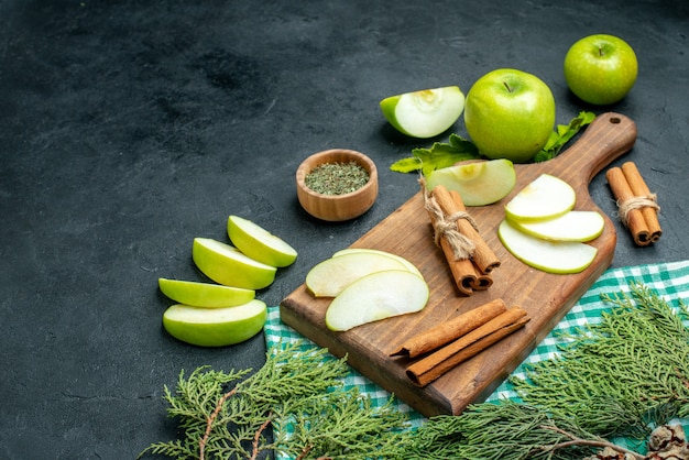 Bottom view apple slices and cinnamons on wood board dried mint powder in bowl apples green and white checkered tablecloth pine tree branches on black ground free place