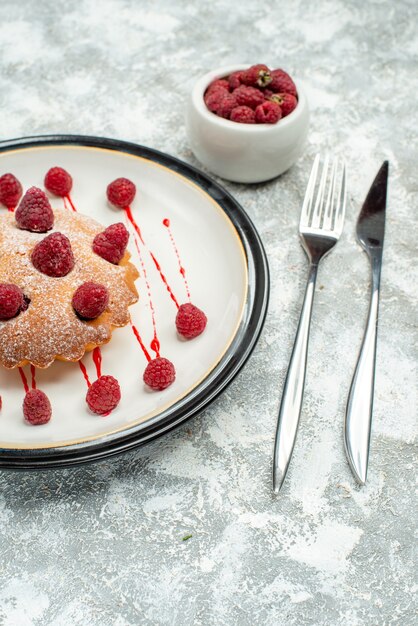 Bottom view berry cake on white oval plate raspberries in bowl fork and dinner knife on grey surface