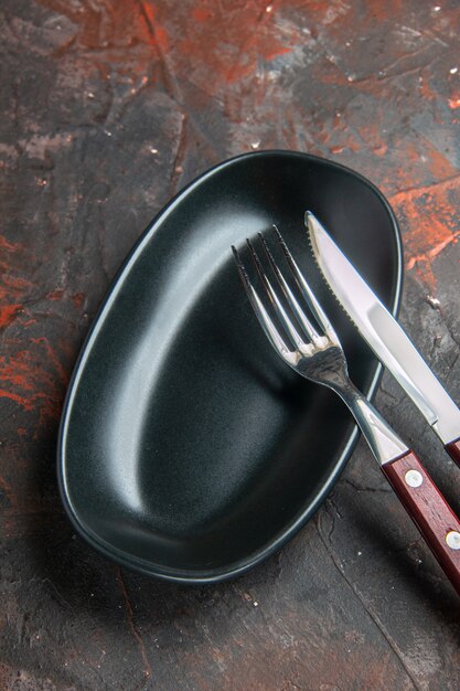 Bottom view black oval plate knife and fork on dark red table copy space