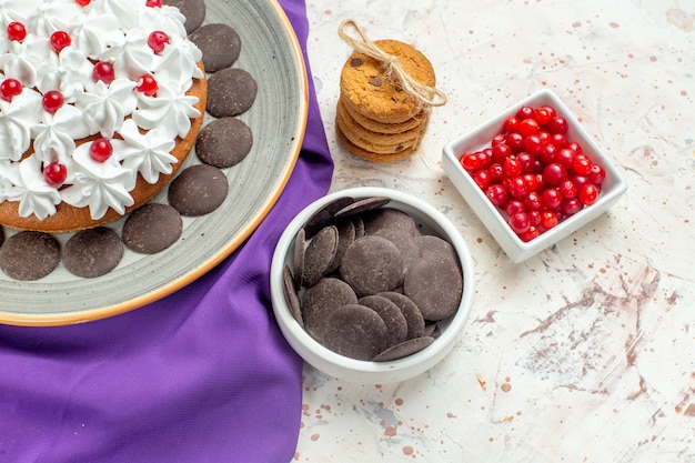 Bottom view cake with pastry cream on grey plate purple shawl cookies tied with rope berries and chocolate in bowl on white table