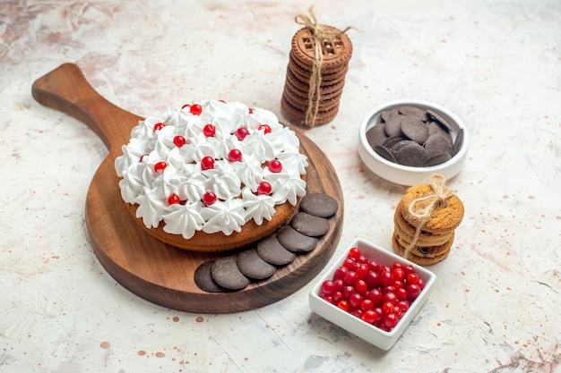 Bottom view cake with white cream on wooden cutting board bowls with berries and chocolate cookies tied with rope on light grey table