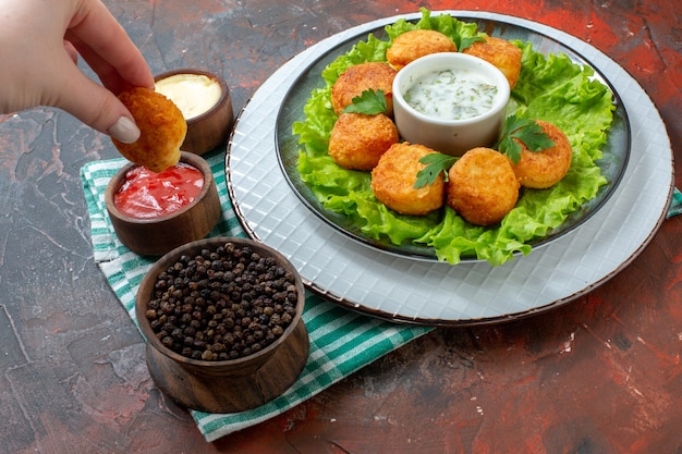 Bottom view chicken nuggets lettuce and sauce on plate black pepper in bowl sauces in small bowls nugget in female hand on dark table