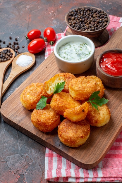 Bottom view chicken nuggets on wood board with sauces cherry tomatoes wooden spoons black pepper in bowl on dark background