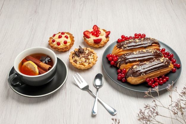 Free photo bottom view chocolate eclairs and currants on the grey plate cookies lemon cinnamon tea spoon and fork in diagonal vector on the white wooden table