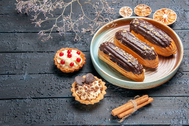 Free photo bottom view chocolate eclairs on oval plate dried flower branch tarts cinnamon and dried oranges on the dark wooden table with copy space