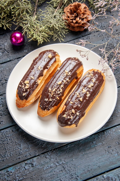 Bottom view chocolate eclairs on white oval plate fir-tree branches cone christmas toy on dark wooden table