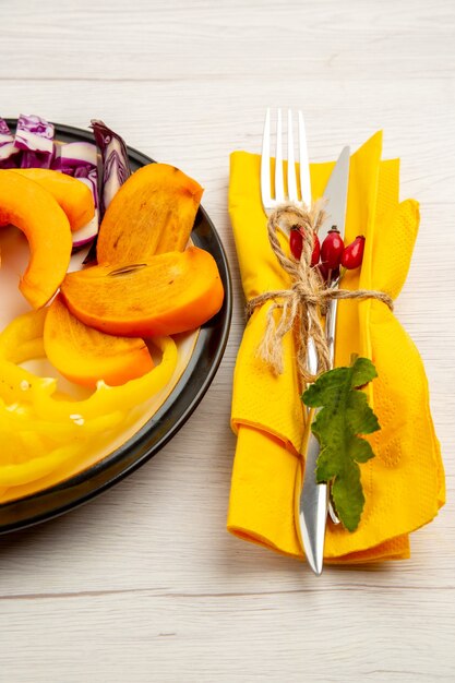 Bottom view chopped vegetables and fruits pumpkin bell peppers persimmon on black plate fork and knife on yellow napkin on white surface