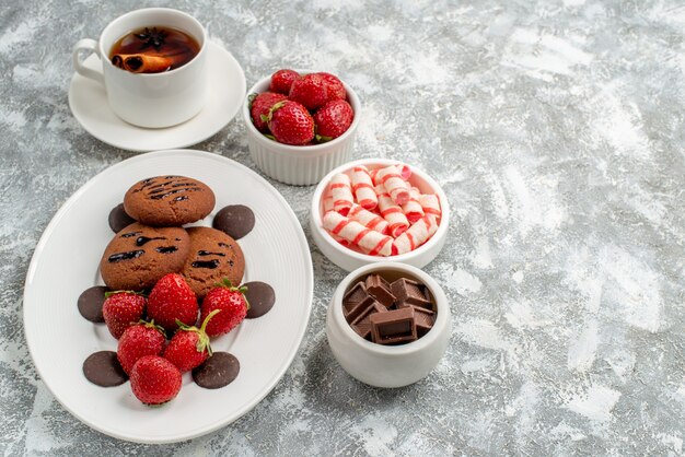 Bottom view cookies strawberries and round chocolates on the oval plate bowls of candies strawberries chocolates cinnamon anise tea at the left side of the grey-white table