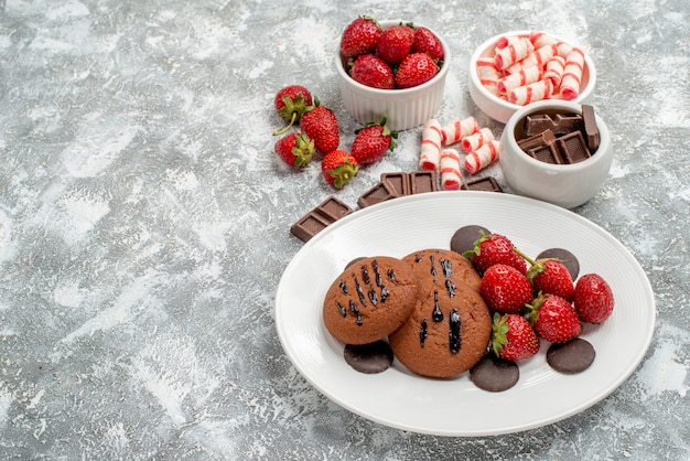 Bottom view cookies strawberries and round chocolates on the white plate and bowls of candies strawberries chocolates on the grey-white ground