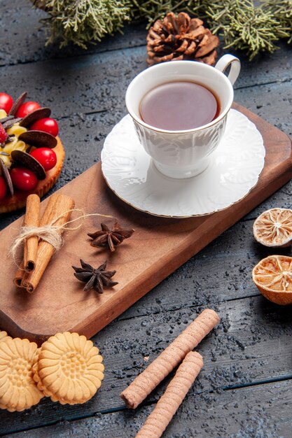 Bottom view a cup of tea anise seeds and cinnamon on wooden serving plate pinecone berry cake dried oranges and different cookies on dark wooden background