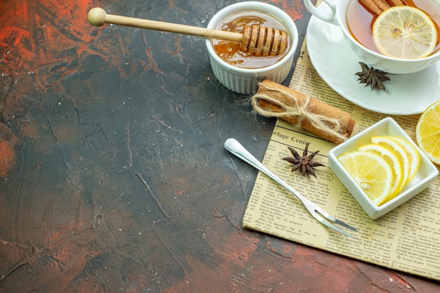 Free photo bottom view cup of tea lemon slices in small bowl fork cinnamon sticks anises honey and honey stick in bowl on newspaper notepad on dark red table with copy place