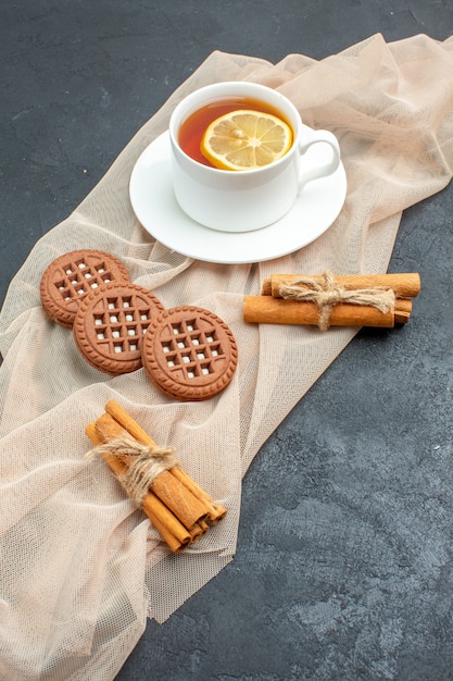 Bottom view a cup of tea with lemon cinnamon sticks cookies on beige shawl on dark surface