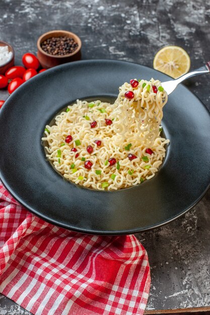 Bottom view ramen noodles raised on fork on black plate black pepper in bowl cut lemon cherry tomatoes on dark table