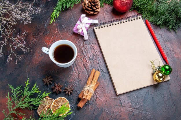 Bottom view red pen a notebook pine tree branches xmas tree ball toys cinnamon sticks a cup of tea on dark red background